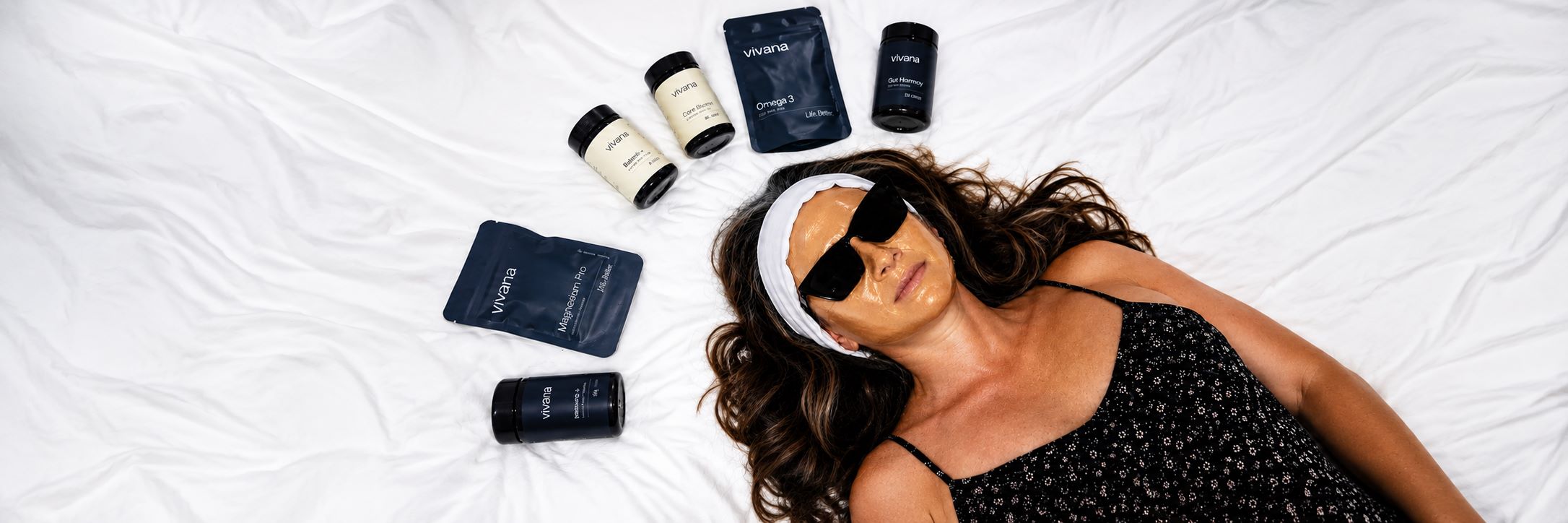 A woman lies on white bedding wearing a black patterned top, sunglasses, and a white headband, surrounded by several skincare or beauty products including bottles and pouches.