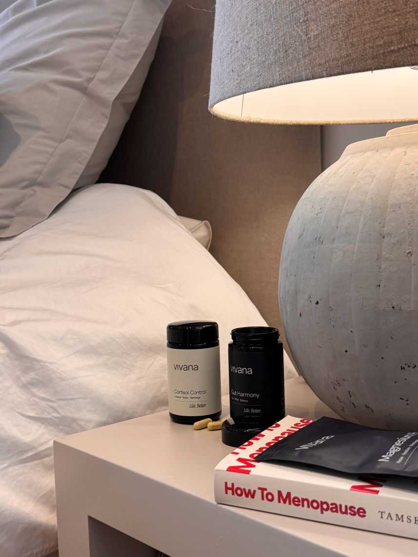 Two black supplement bottles from the brand Vivana, displayed on a bedside table next to a lamp and a book titled 'How To Menopause'.