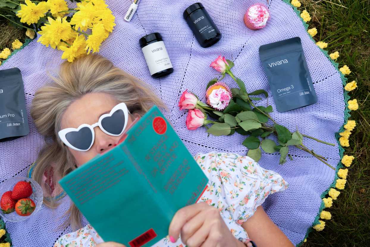 A woman wearing heart-shaped white sunglasses lies on a purple picnic blanket outdoors, reading a teal book. Surrounding her are yellow flowers, pink roses, strawberries, two Vivana supplement bottles