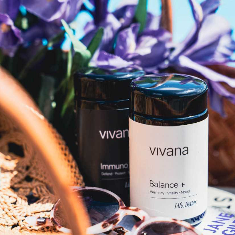 Two jars of vivana supplements, one black and one white, styled in a wicker basket with sunglasses, a straw hat, and purple flowers.