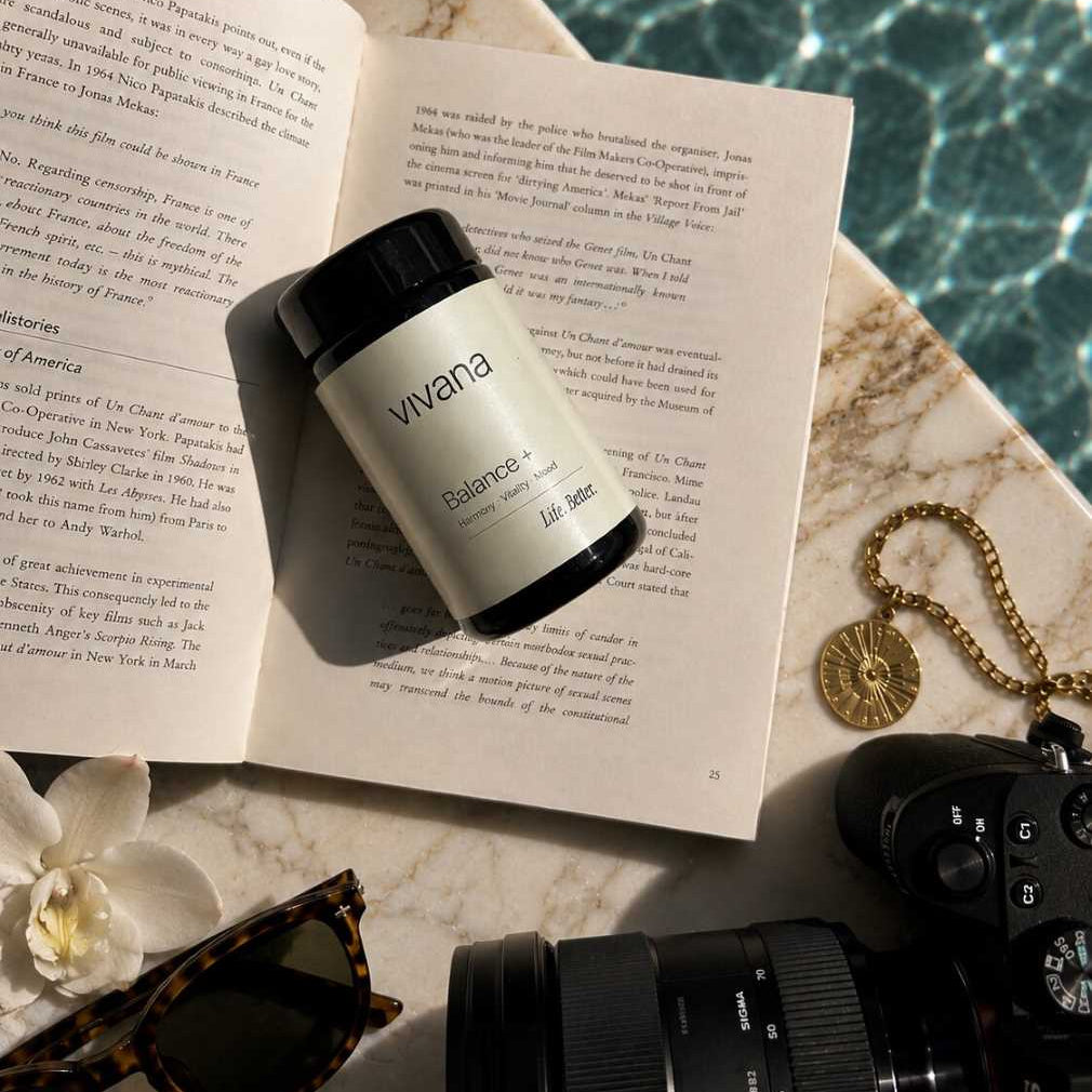 A bottle of Vivana Balance+ with Safran as Safr'Inside lies on an open book, styled with a camera, sunglasses, a necklace, and a flower on a marble surface near water
