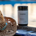 A jar of vivana Balance + placed on a book outdoors, with a woven straw hat in the foreground and a blurred blue-toned background.