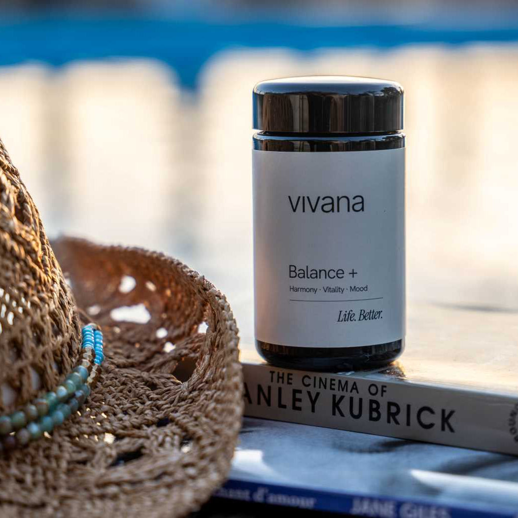 A jar of vivana Balance + placed on a book outdoors, with a woven straw hat in the foreground and a blurred blue-toned background.