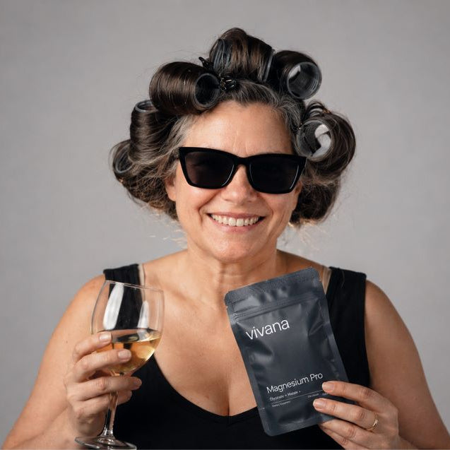 A smiling woman with dark hair in curlers and black sunglasses holds a black stand-up pouch labeled 'vivana Magnesium Pro' and a glass of white wine against a plain gray studio background.