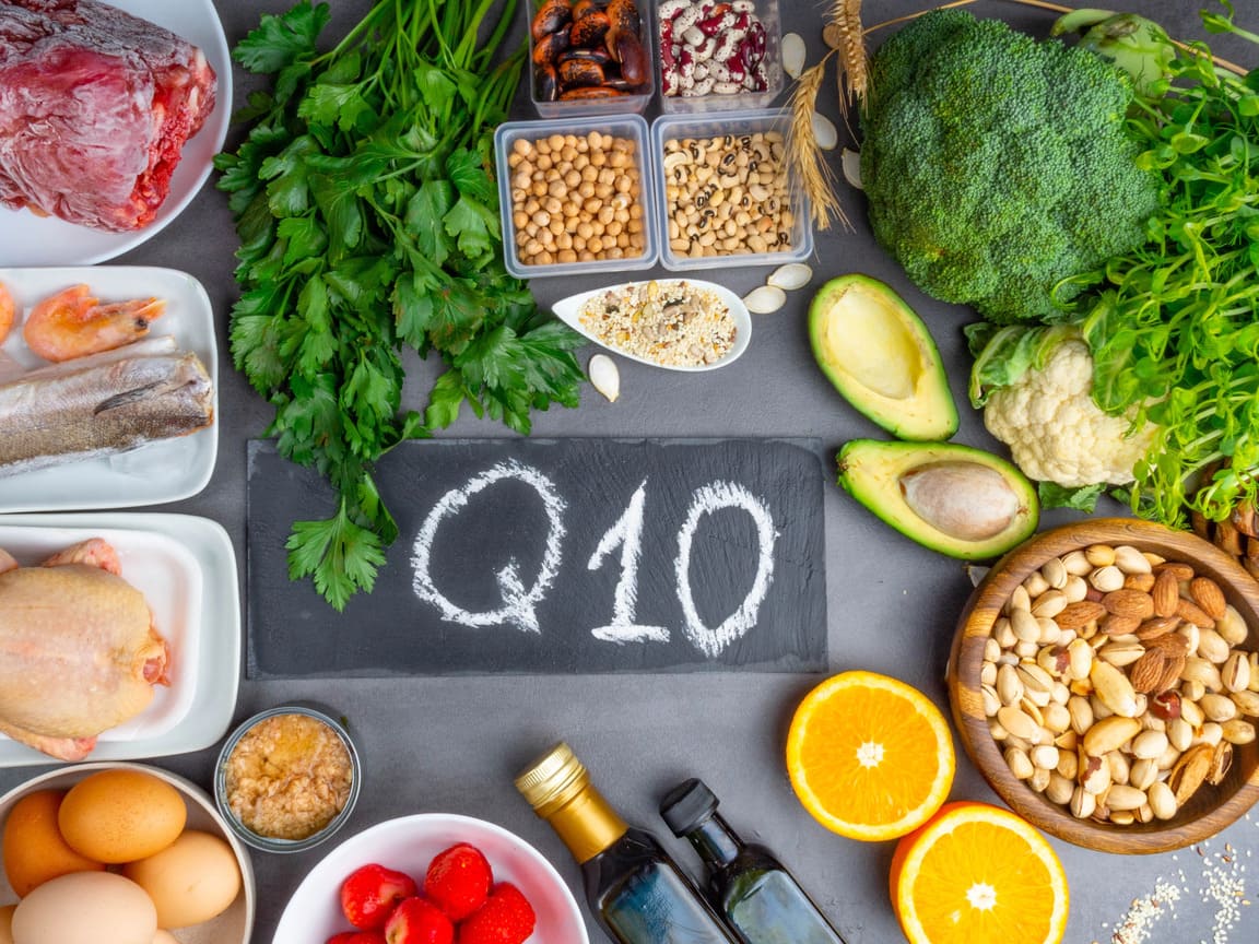 A styled flat-lay of various foods arranged around a central black slate sign with the chalk-written text 'Q10'. The foods include broccoli, avocado, nuts, eggs, tomatoes, herbs, fish, meat, and bottles of oil on a dark gray surface.