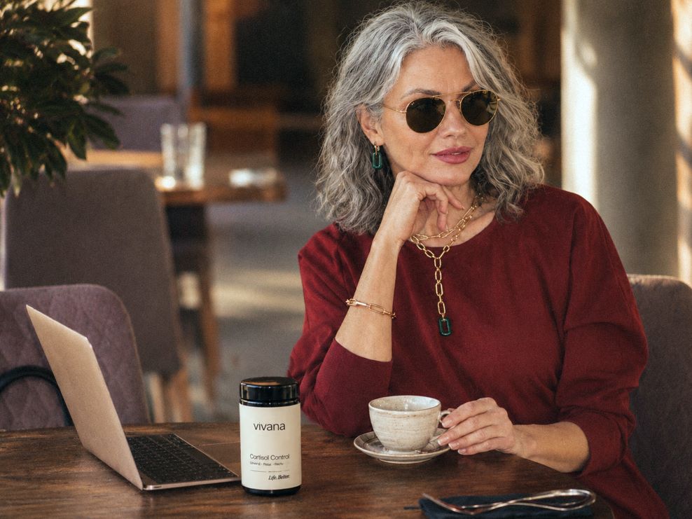 A woman with gray hair and round sunglasses sits at a cafe table wearing a burgundy top, holding a teacup. On the table in front of her is a jar of Vivana Cortisol Control.
