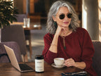 A woman with gray hair and round sunglasses sits at a cafe table wearing a burgundy top, holding a teacup. On the table in front of her is a jar of Vivana Cortisol Control.