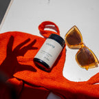 A white jar of vivana Cortisol Control stree support with Ashwagandha KSM-66 with a black lid, styled on an orange textured towel next to yellow sunglasses on a white surface.