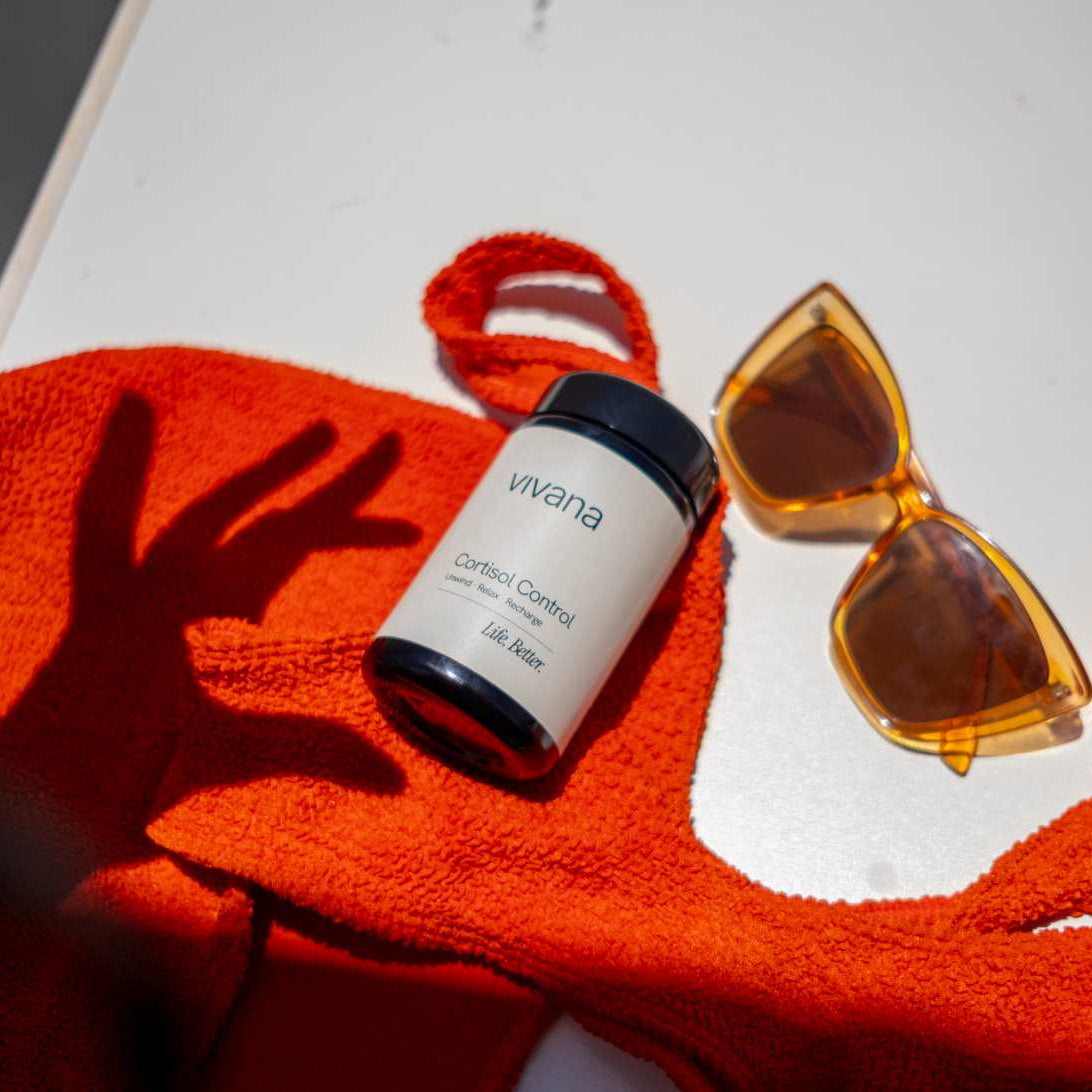 A white jar of vivana Cortisol Control stree support with Ashwagandha KSM-66 with a black lid, styled on an orange textured towel next to yellow sunglasses on a white surface.