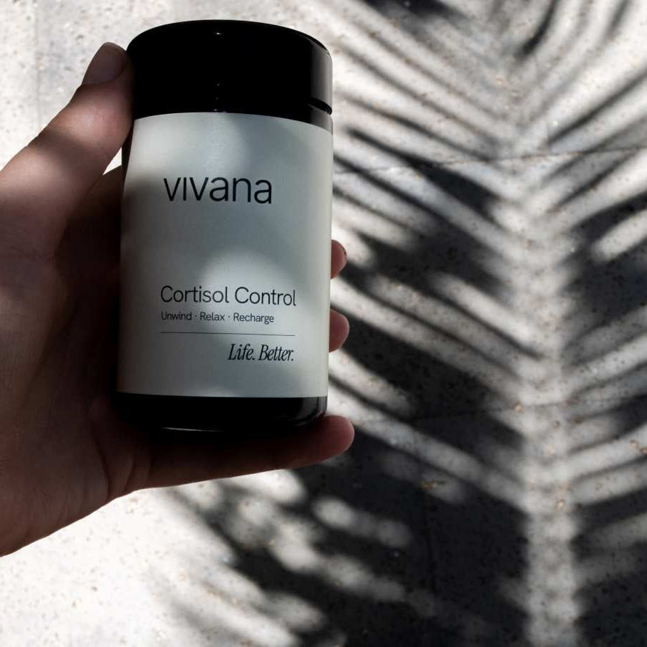 A hand holding a white jar of vivana Cortisol Control with black lid, set against a concrete background with palm leaf shadows.