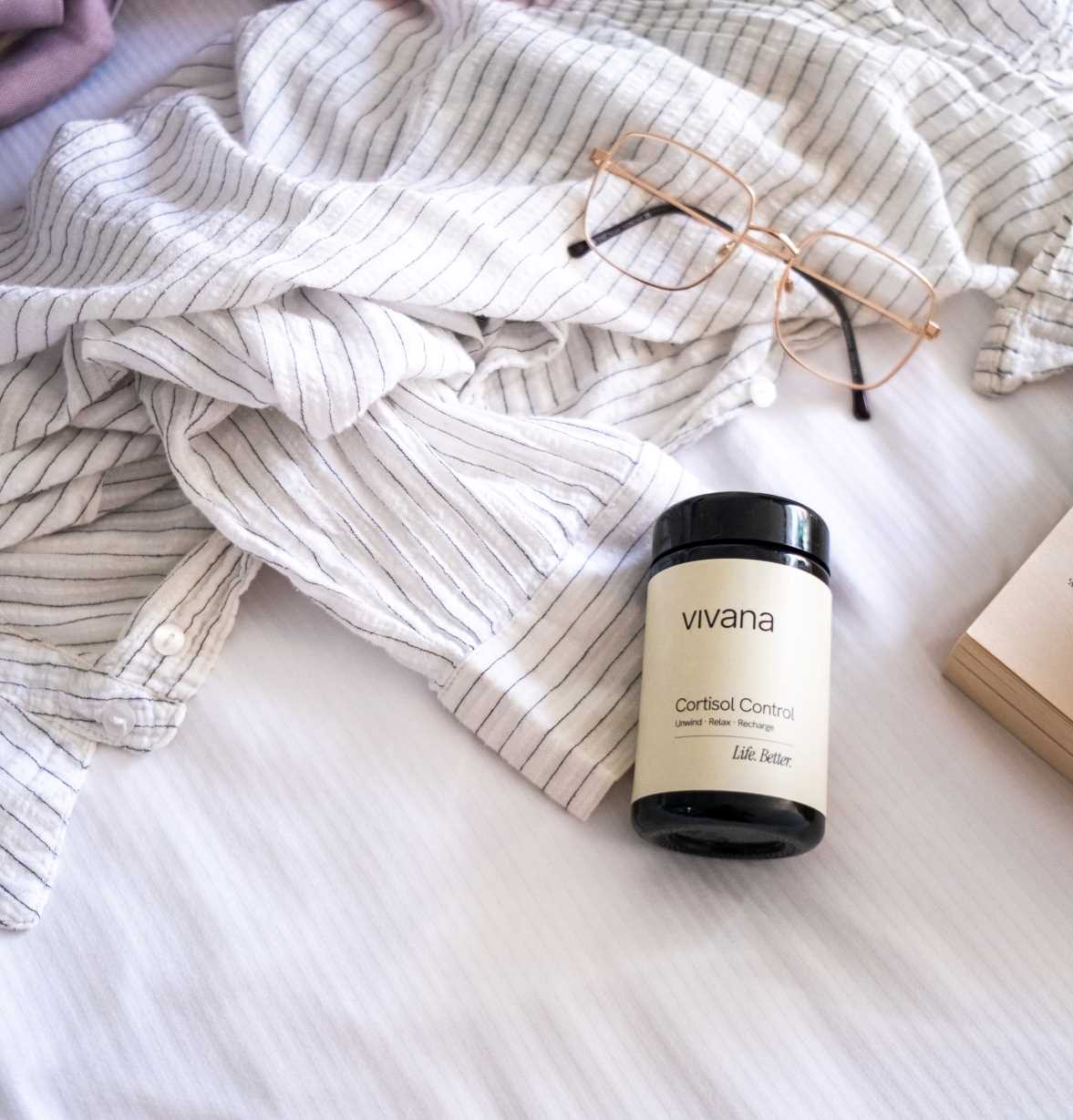 A jar of vivana Cortisol Control sleep and stress support on a white bed with a striped shirt, gold-framed glasses, and a book.