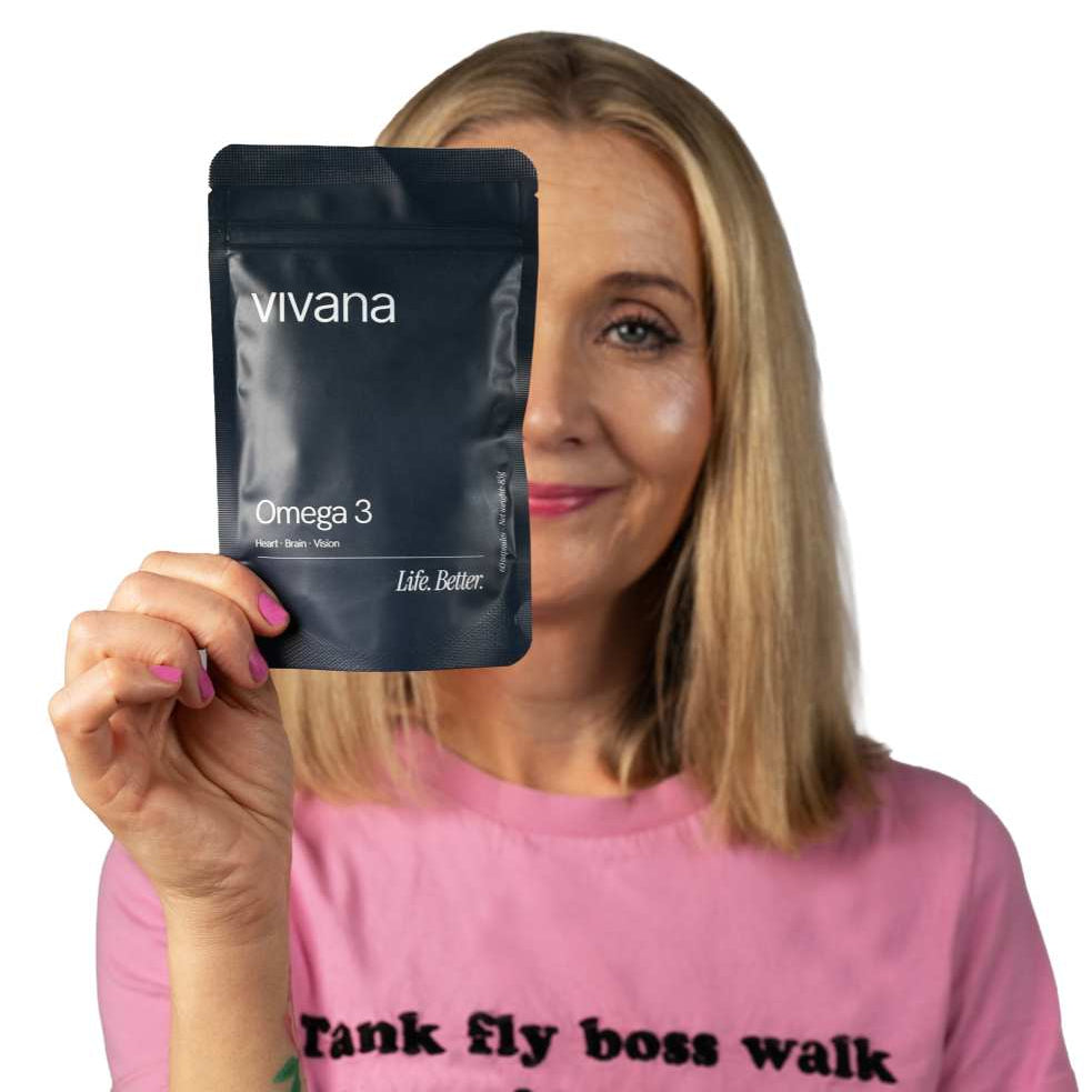 A woman with blonde hair holds up a black stand-up pouch of 'vivana Omega 3' fish oil. She is wearing a pink t-shirt with black text that reads 'tank fly boss walk jam nitty-gritty' against a solid white background.