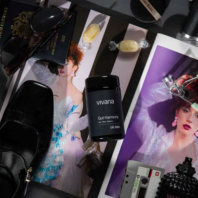 A styled flat-lay featuring a black jar of vivana Gut-Harmony in the center, surrounded by fashion props including black leather shoes, sunglasses, a camera, and printed fashion magazines on a dark surface.