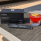 Vivana Collagen Pro supplement packaging shown outdoors on a woven table mat, with a black product box, two single-serve packets, and a glass of red cocktail with a lemon slice.