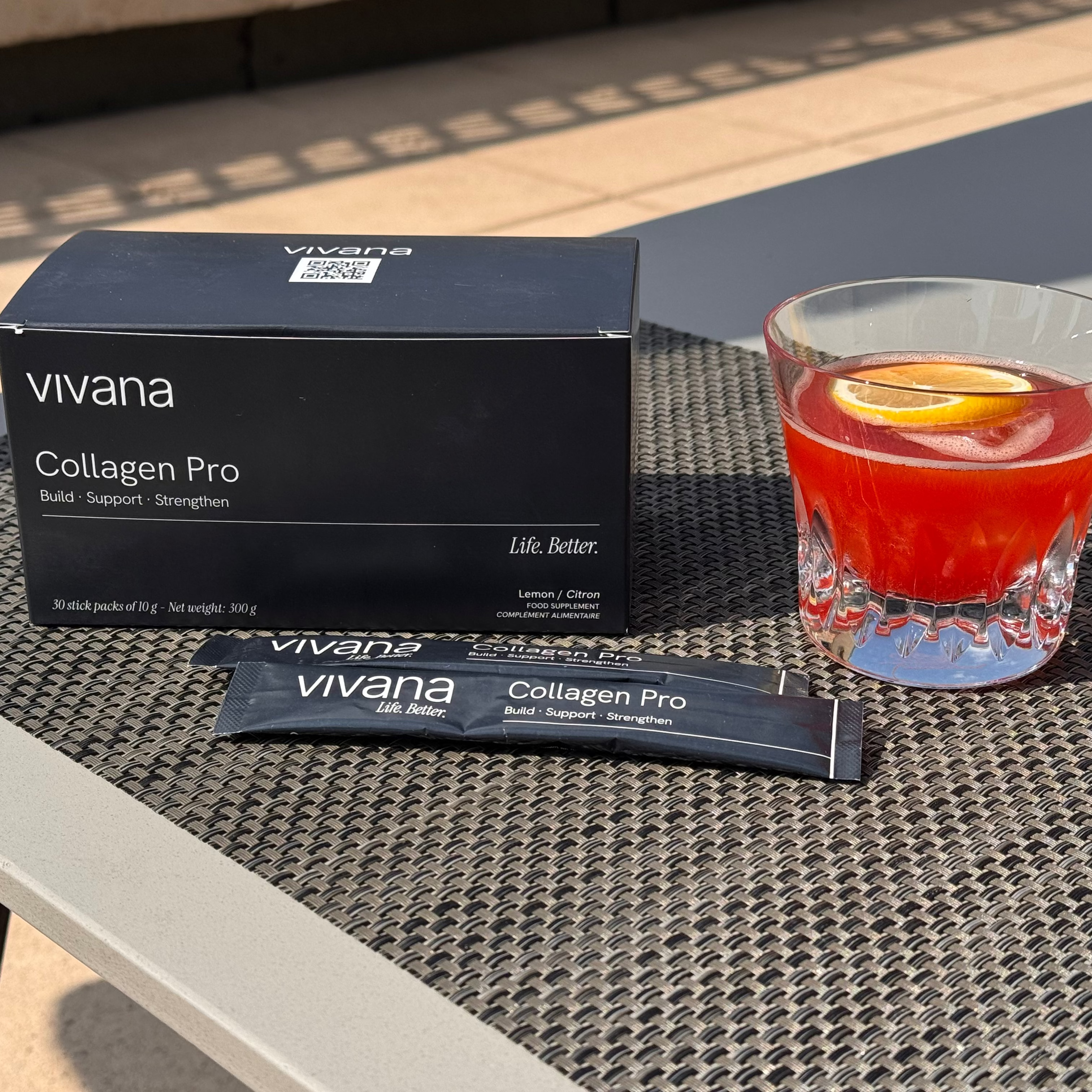 Vivana Collagen Pro supplement packaging shown outdoors on a woven table mat, with a black product box, two single-serve packets, and a glass of red cocktail with a lemon slice.