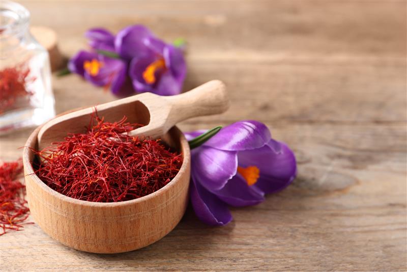 A wooden bowl filled with bright red Safr'Inside saffron threads and a small wooden scoop, styled on a rustic wooden surface with purple crocuses and a glass jar of saffron in the background.